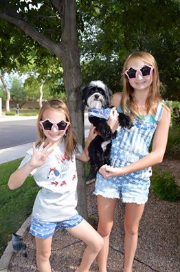 a girl with star-shaped sunglasses next to a taller girl holding a black dog and wearing star-shaped sunglasses